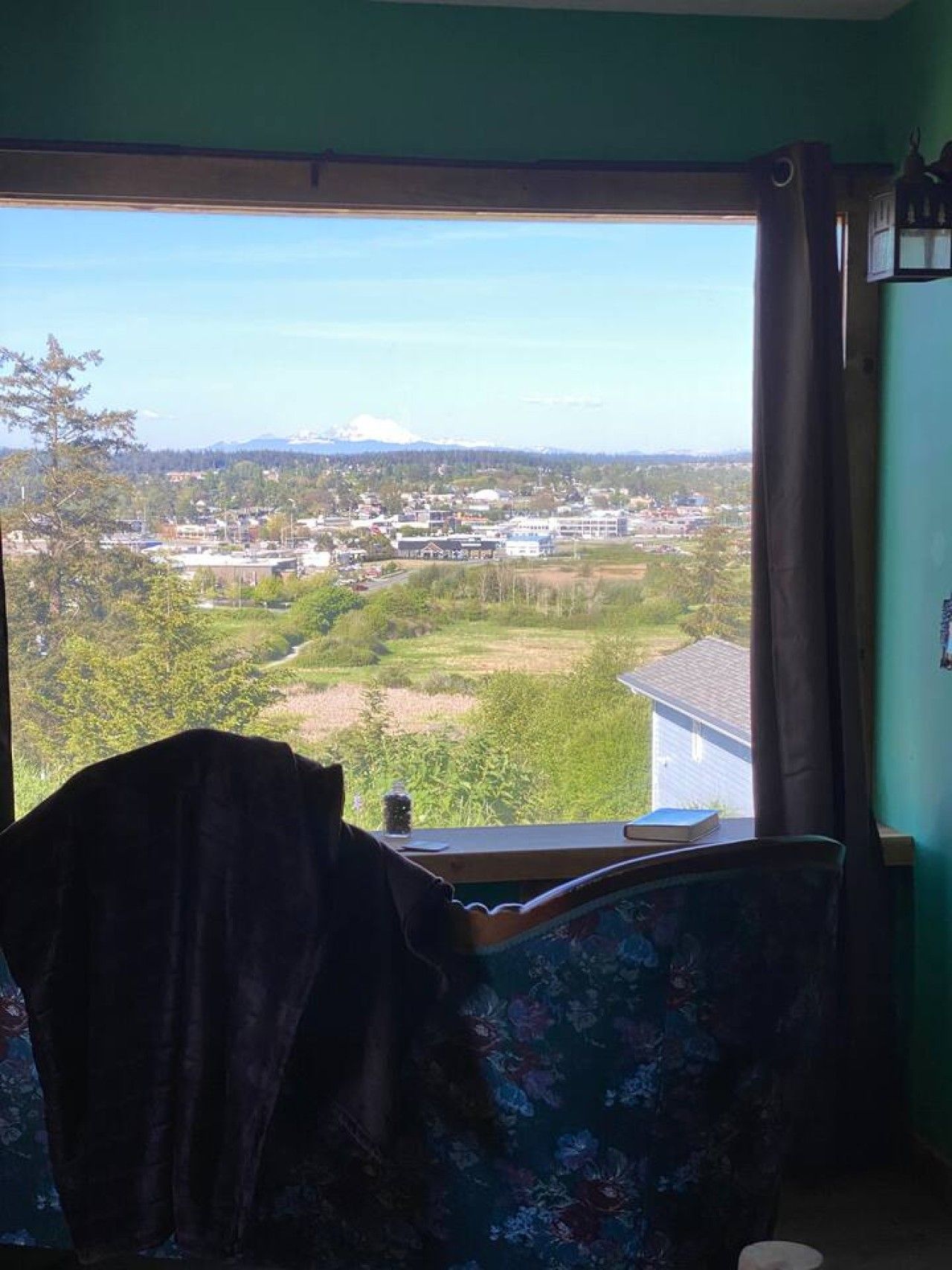 A room with a view of a town and mountains, a chair, and a table with a book and a bottle.
