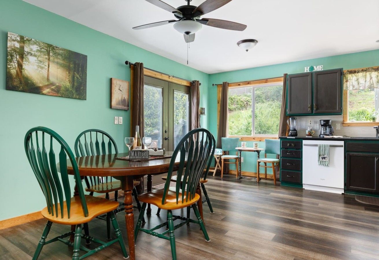 A dining area with a wooden table and chairs, a ceiling fan, and a kitchen with cabinets and a window.