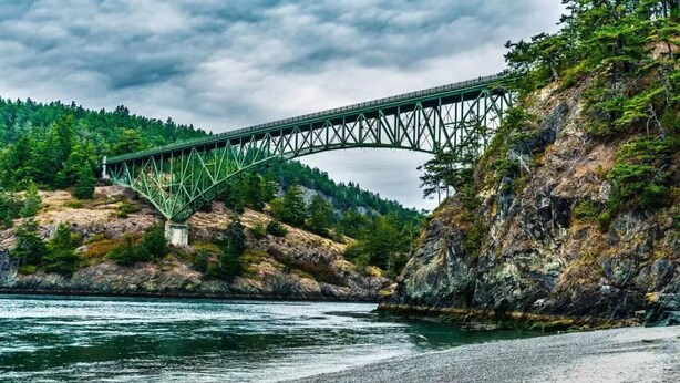 Un pont d’acier enjambant une rivière, avec des berges rocheuses et des collines boisées.
