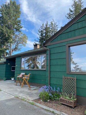 A green building with large windows, a wooden deck with chairs, and a garden with flowers. - Cabin * Hot tub * Fire pit * View * Getaway! (Oak Harbor)