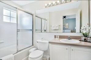A modern bathroom with a glass shower enclosure, a white toilet, a sink with a vanity, and a large mirror with lights above it. - Central to all the fun Disney (Kissimmee)