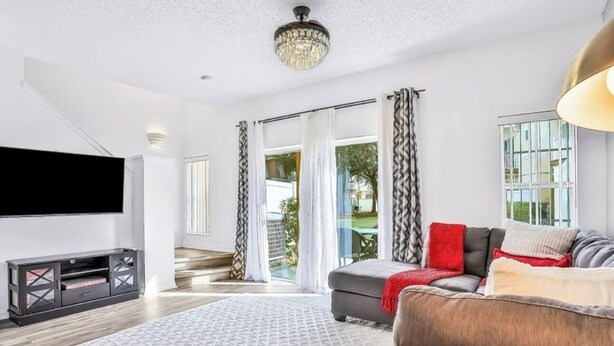 A modern living room with a grey sofa, a patterned rug, a flat-screen TV, and a chandelier. - Central to all the fun Disney (Kissimmee)