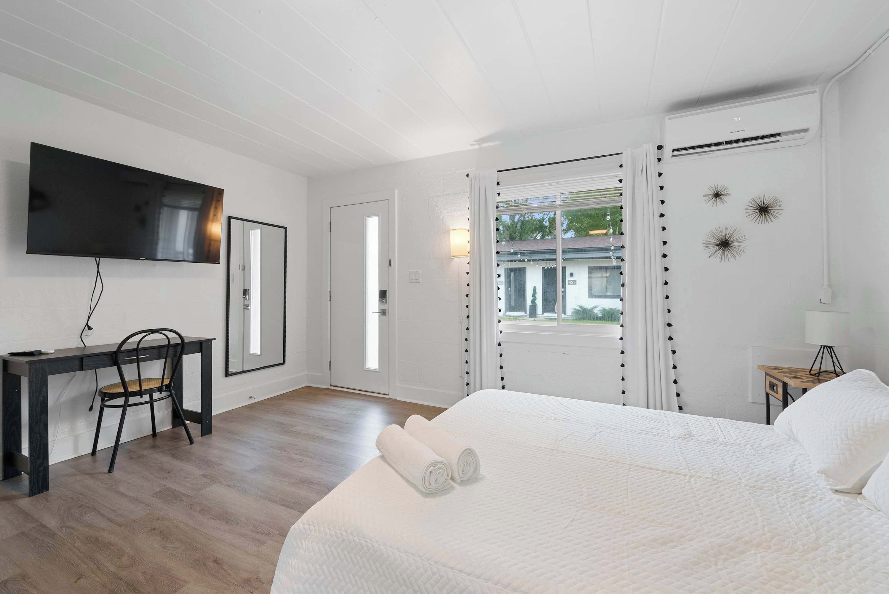 A bedroom with a large bed, a desk with a chair, a flat-screen TV, and a window with curtains.
