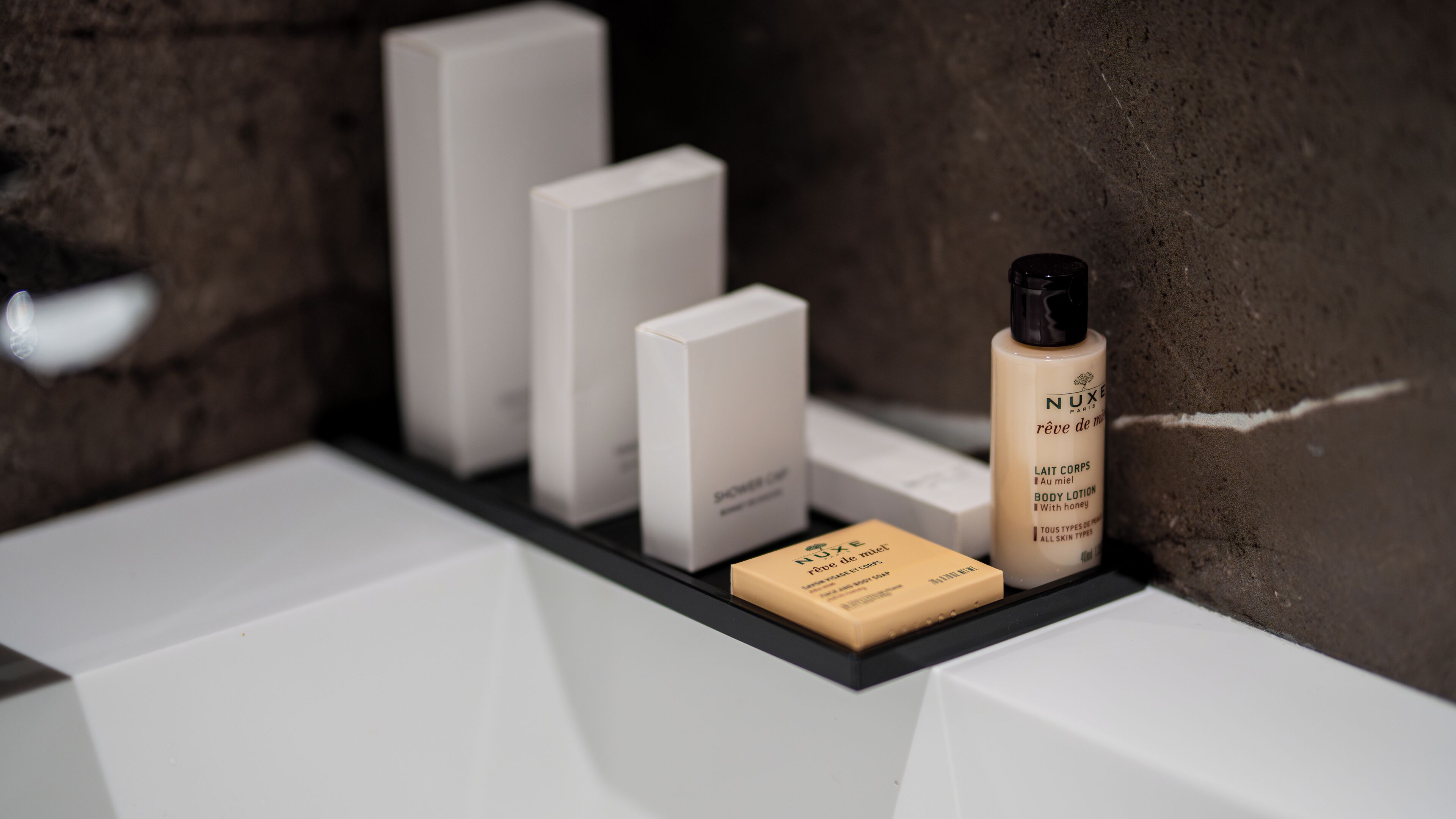 A bathroom shelf with a bottle of Nuxe body lotion, a soap bar, and two white boxes.