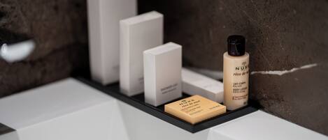 A bathroom shelf with a bottle of Nuxe body lotion, a soap bar, and two white boxes.