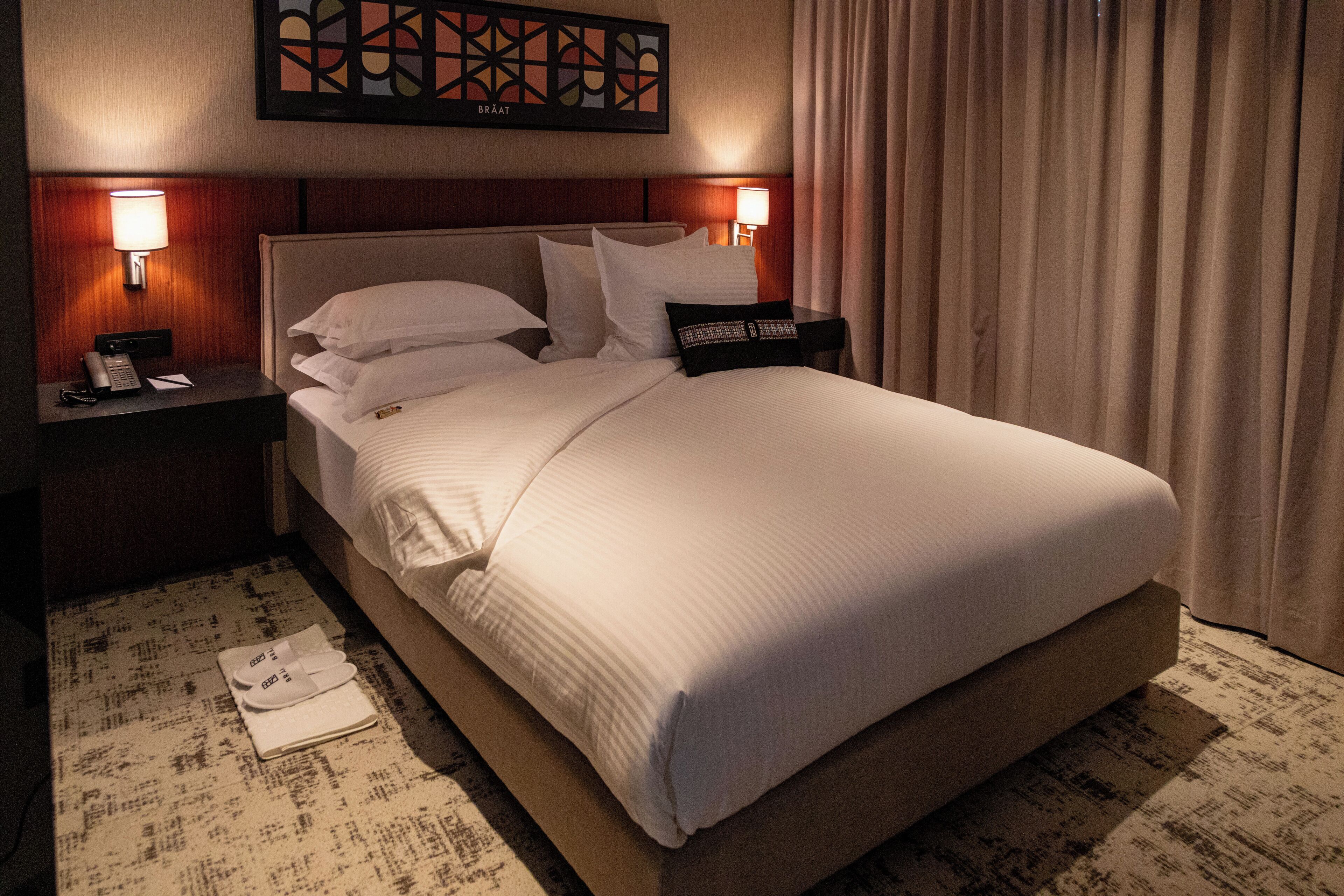 A hotel room with a large bed, two bedside lamps, a nightstand with a phone, and a wall art piece above the bed.