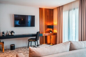 Una habitación de hotel moderna con una televisión de pantalla plana montada en la pared, un escritorio con una silla, un sofá y un amplio ventanal con cortinas.