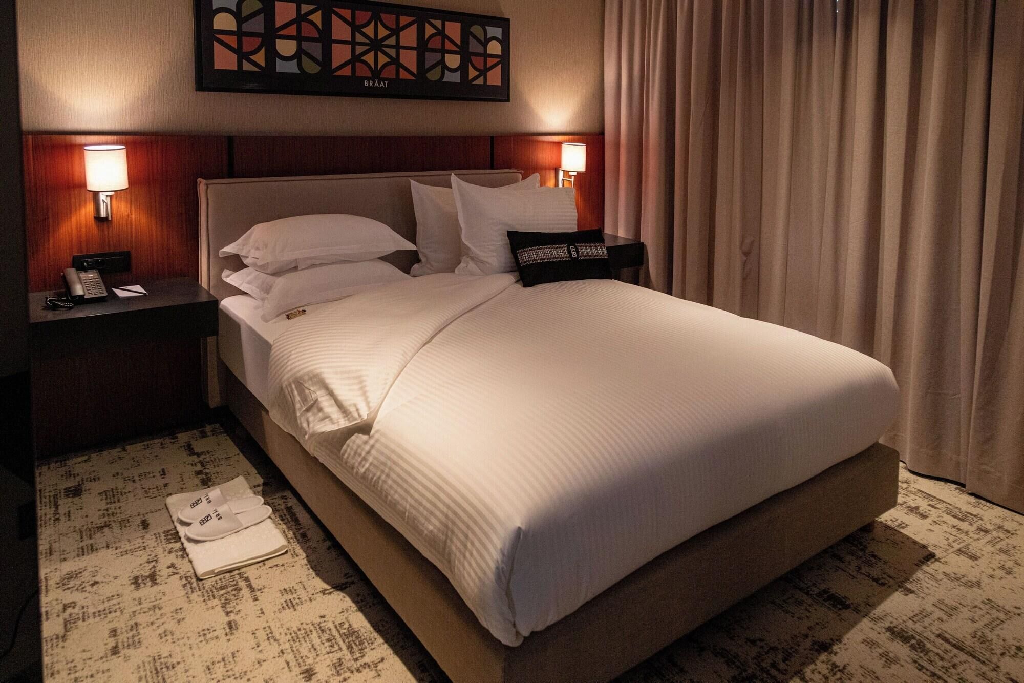 Une chambre d’hôtel avec un grand lit, deux tables de chevet avec des lampes, une œuvre d’art accrochée au mur et un rideau.