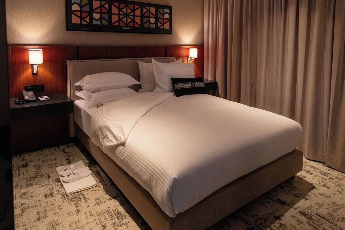 Una habitación de hotel con una cama grande, dos mesitas de noche con lámparas, una obra de arte colgada en la pared y una cortina.