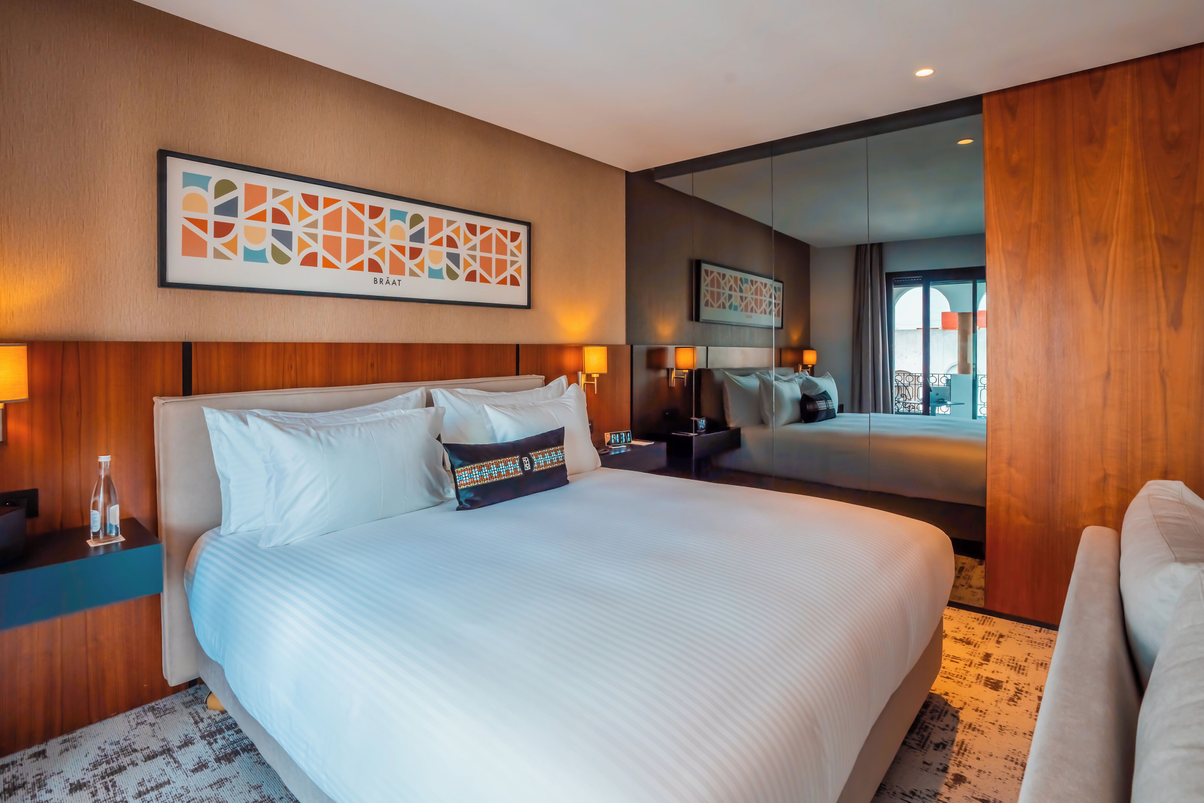 A hotel room with a large bed, a wooden headboard, a nightstand with a lamp, and a large mirror.