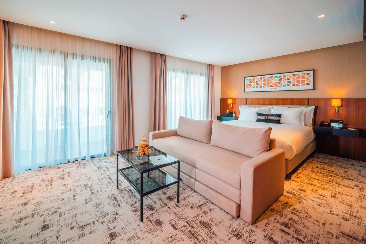 Una habitación de hotel con una cama, un sofá, una mesa de centro de vidrio y un ventanal con cortinas.