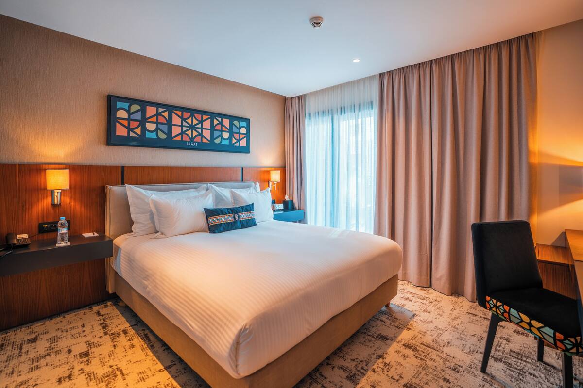 Habitación de hotel con una cama grande, dos lámparas de noche, un adorno en la pared, una silla y un ventanal con cortinas.