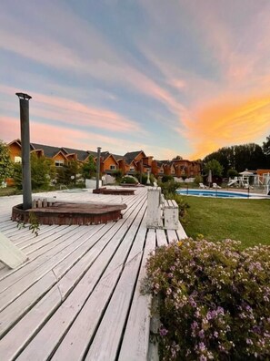 A wooden deck with a barbecue area, a pool, and a row of cabins with a sunset in the background. - APART HOTEL SOL Y LAGO (Villarrica)