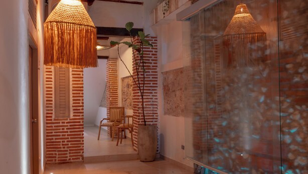 A hallway with a brick wall, wooden shutters, a large potted plant, and a glass partition with a water feature. - Casa Helena Hostel Cartagena (Cartagena)
