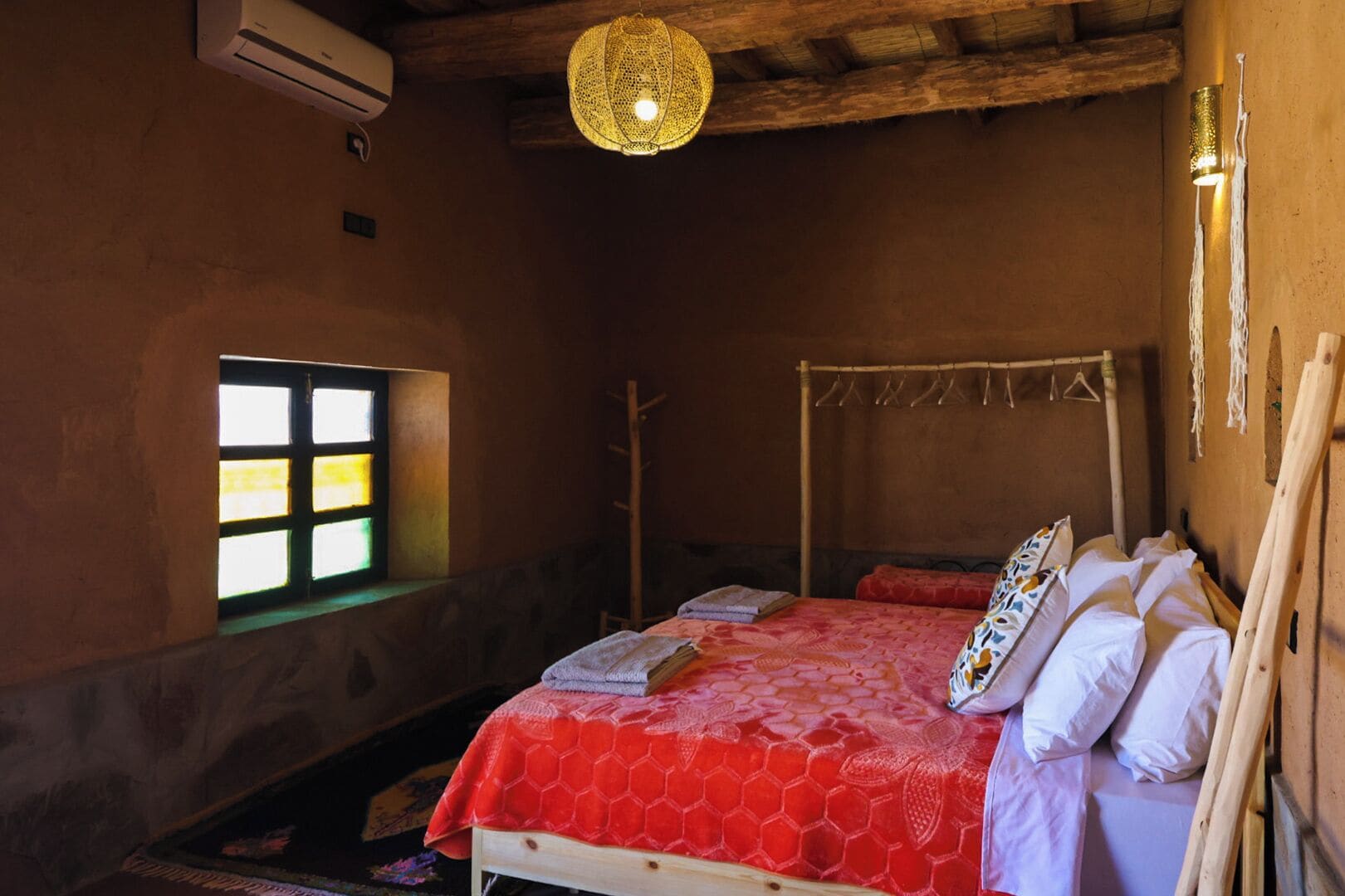 A bedroom with a wooden bed, a window with a view, a hanging light fixture, and a wall-mounted air conditioner.