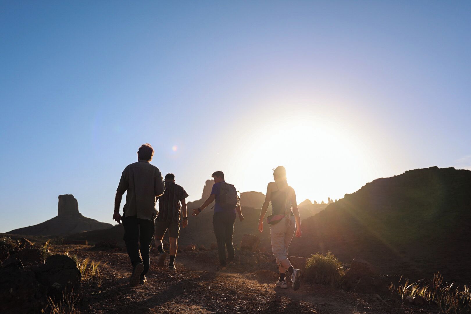 A group of people hiking on a dirt trail with a clear sky and a large rock formation in the background.