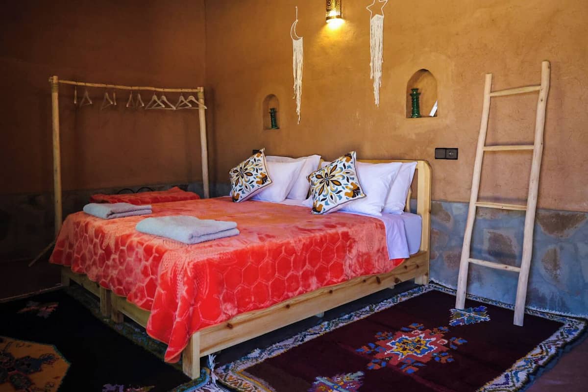 A single bed with a red and white patterned bedspread, two pillows with floral designs, a wooden ladder against the wall, and a hanging light fixture.