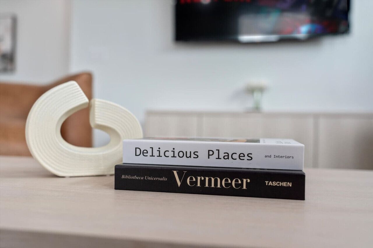 A stack of books titled 'Delicious Places and Interiors' and 'Vermeer' on a table with a roll of toilet paper beside them.