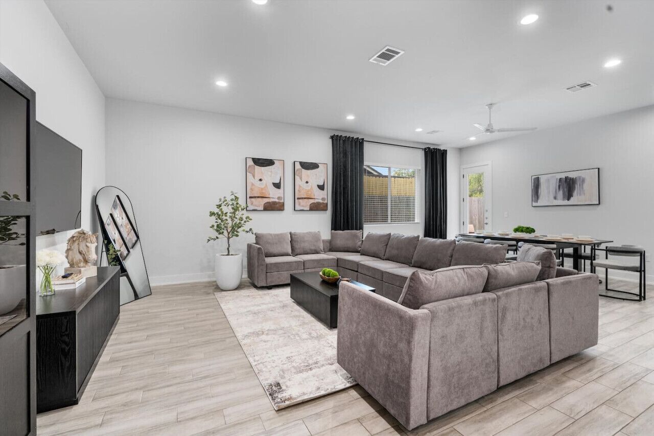 A modern living room with a grey sectional sofa, a coffee table, and a flat-screen TV on a black stand. There is a dining area with a table and chairs, and a potted plant in the corner.