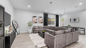 Un salon moderne avec un canapé d’angle gris, une table basse et un téléviseur à écran plat posé sur un meuble noir. On y trouve également un coin repas avec une table et des chaises, ainsi qu’une plante en pot dans un coin.