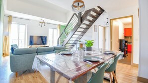 A dining area with a wooden table, clear plastic tablecloth, and green chairs. A staircase with a metal railing leads to an upper level.