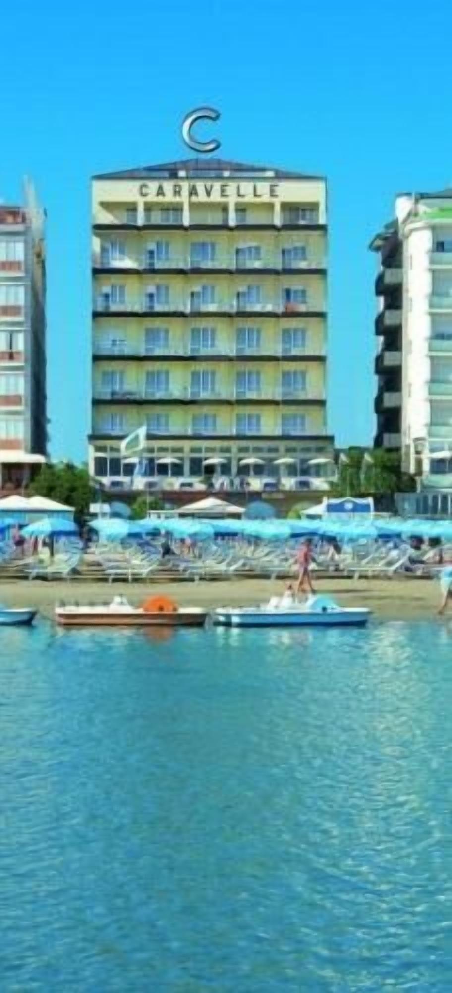 Un hotel llamado CARAVELLE con varios pisos, un área de playa con sombrillas y barcos en el agua.