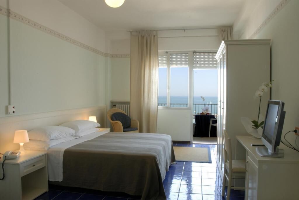 A hotel room with a bed, bedside table with a lamp, a desk with a computer, and a view of the sea through a window.
