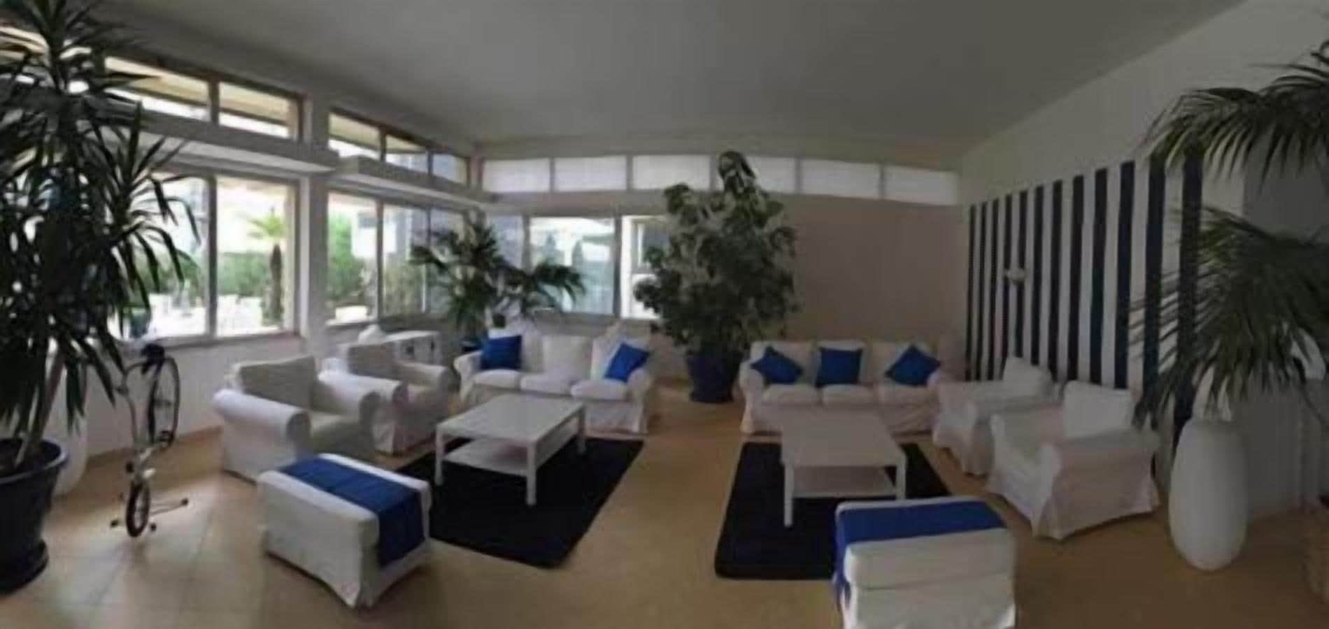 A spacious living area with white furniture, large windows, and potted plants.