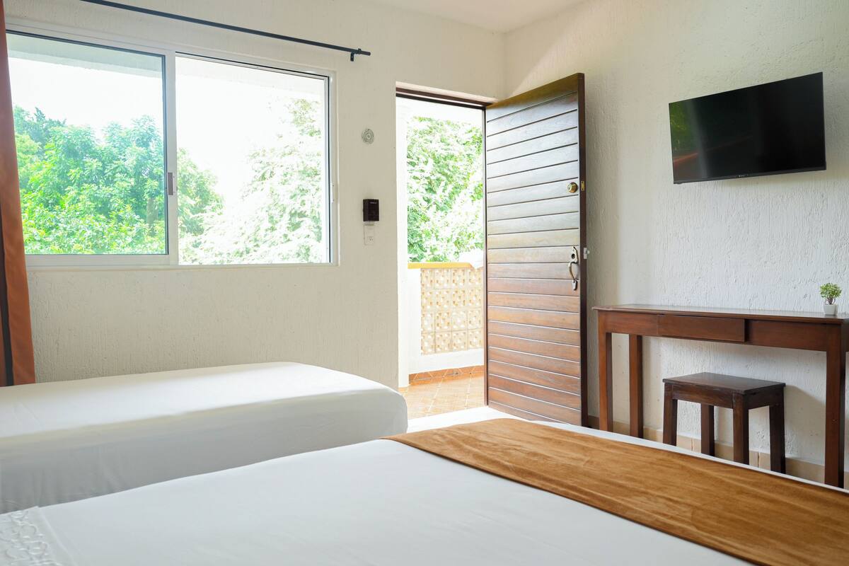 A bedroom with two beds, a TV mounted on the wall, and a view of greenery outside.