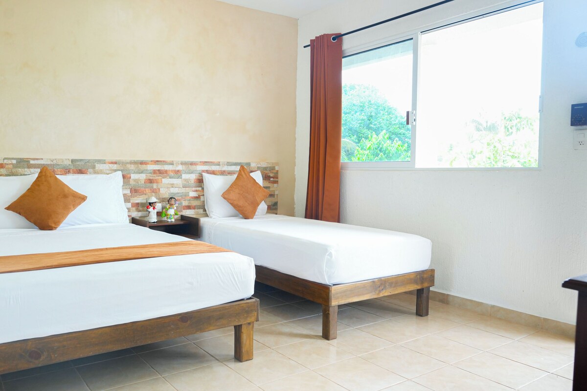 A hotel room with two beds, a brick-patterned headboard, a window with curtains, and a view of greenery outside.