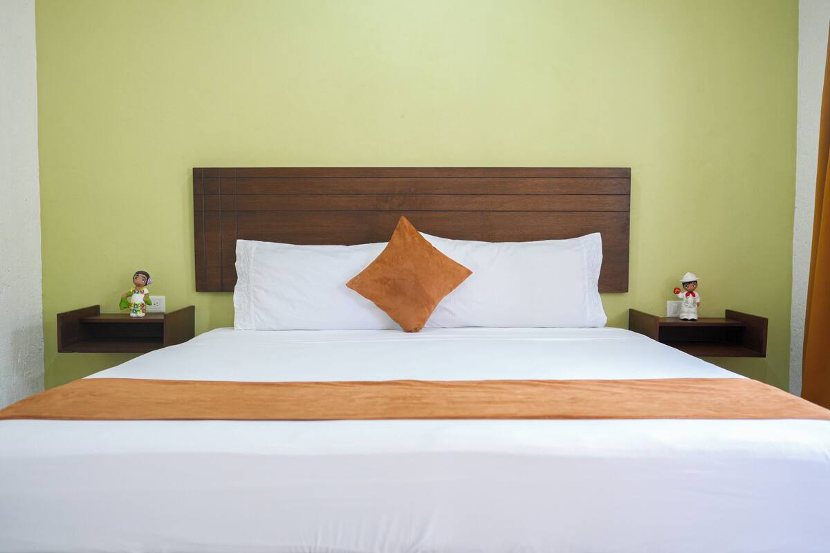A neatly made bed with white linens and a brown accent pillow, flanked by two nightstands with small figurines.