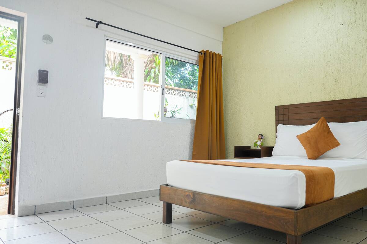 A hotel room with a wooden bed, white bedding, a nightstand with a figurine, a window with curtains, and a view of greenery outside.