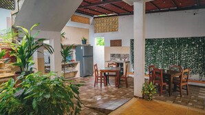A covered patio area with a dining table and chairs, surrounded by potted plants and a kitchen area with a refrigerator.