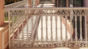 Un balcon doté de balustrades blanches décorées, d’un sol carrelé et offrant une vue sur des palmiers et des bâtiments en arrière-plan.