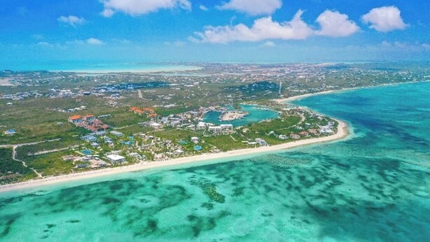 Vue aérienne d’une station balnéaire comprenant plusieurs bâtiments, une plage et des eaux turquoise cristallines.