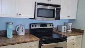 Une cuisine avec des meubles blancs, un four à micro-ondes en acier inoxydable, une cuisinière noire, une machine à café et une bouilloire posées sur le plan de travail.