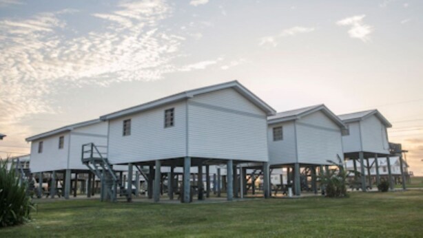 A row of white buildings with a flat roof, supported by pillars, in a grassy area with a clear sky.