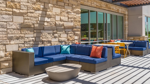 An outdoor patio area with a stone wall, blue and orange upholstered seating, and a round table. - Home2 Suites By Hilton Kearney (Kearney)