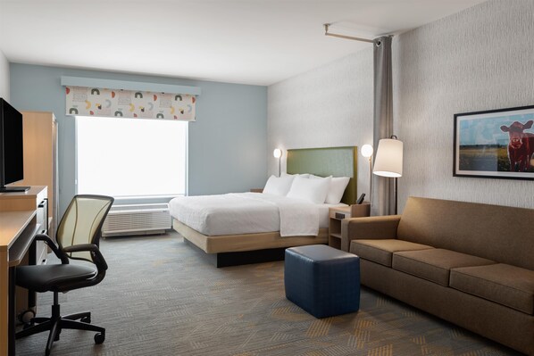 A hotel room with a bed, a desk, a chair, a sofa, and a large window. - Home2 Suites By Hilton Kearney (Kearney)