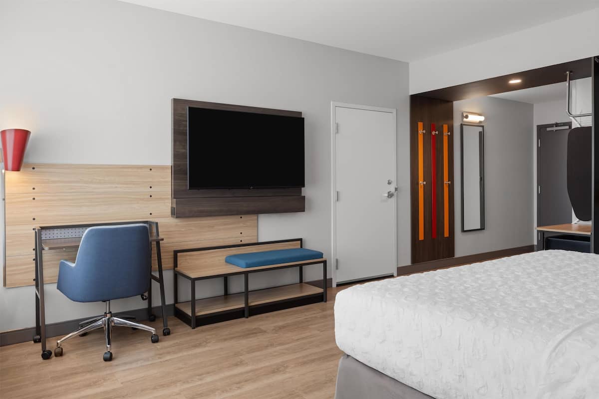 Une chambre d’hôtel comprenant un lit, un bureau avec une chaise, un téléviseur fixé au mur, et une porte agrémentée d’un miroir et de touches de couleur rouge.