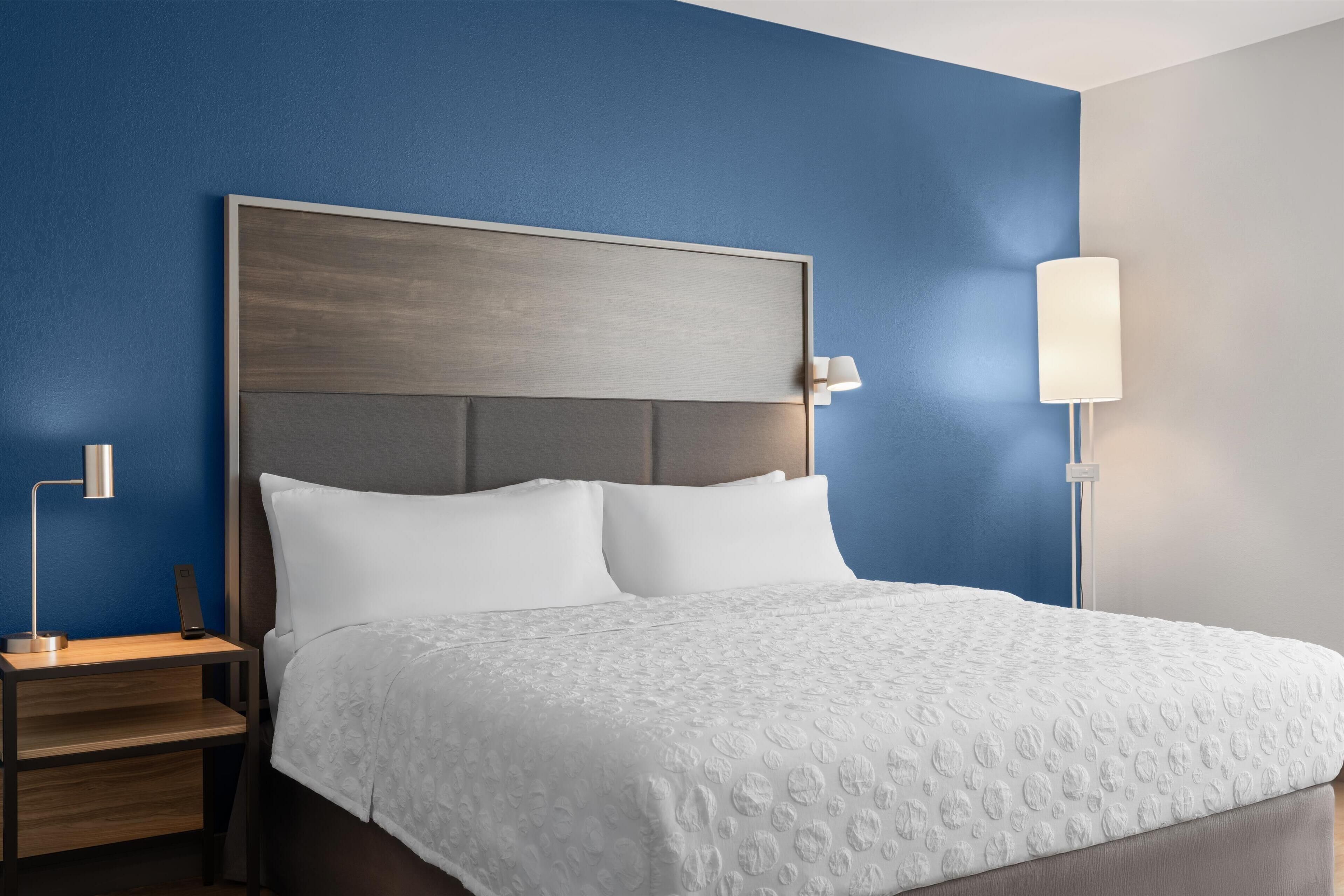 A neatly made bed with a white bedspread and two pillows, a wooden nightstand with a lamp, and a wall lamp on the right side.