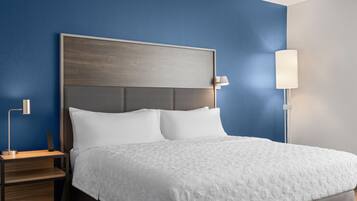 A neatly made bed with a white bedspread and two pillows, a wooden nightstand with a lamp, and a wall lamp on the right side.