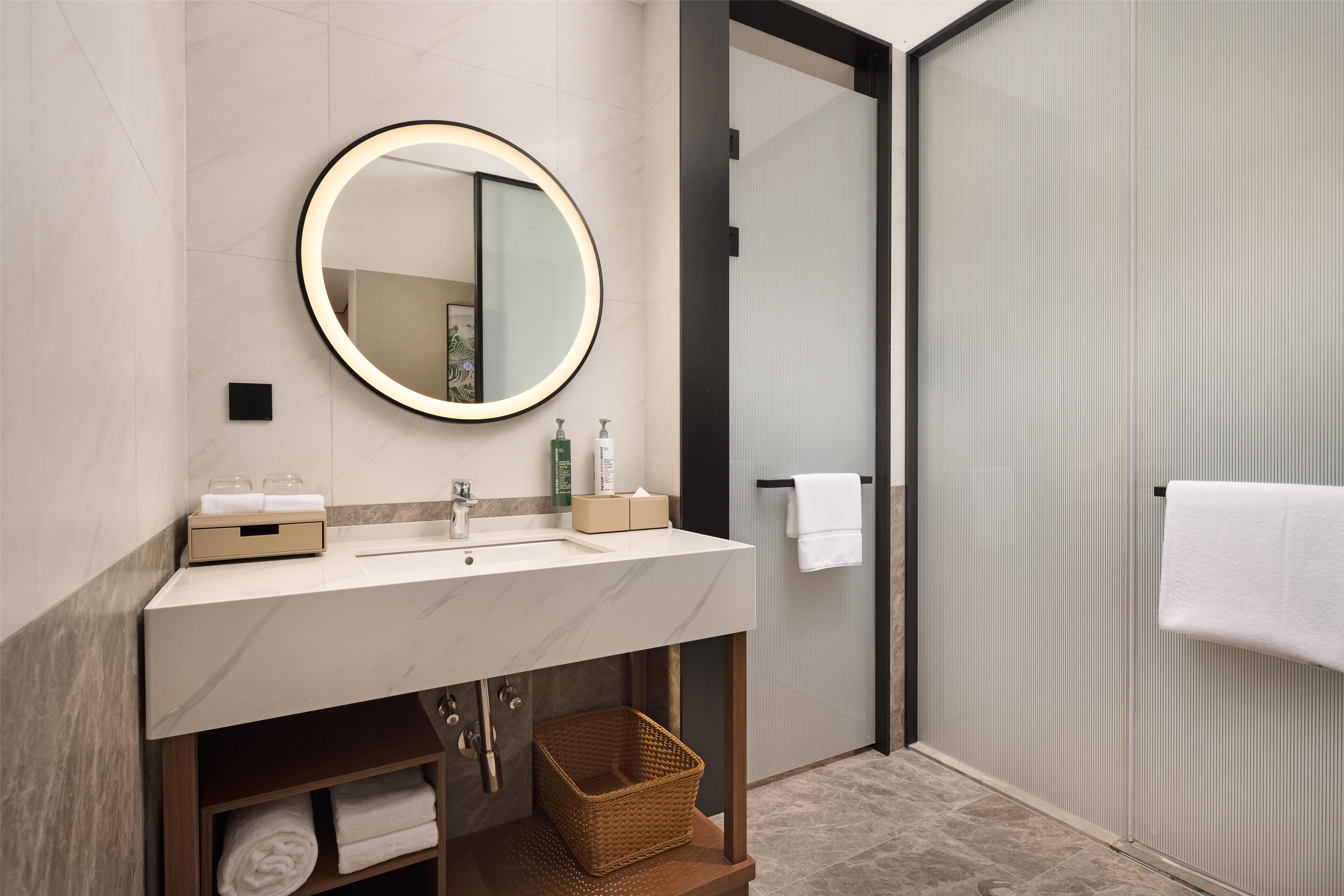 Un baño moderno con lavamanos de mármol, un espejo redondo con luz, un perchero y una cesta debajo del lavamanos.
