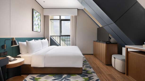 A modern hotel room with a bed, bedside table, lamp, and a view of the city through a large window. - Hilton Garden Inn Guiyang Gui'An New Area (Guiyang)
