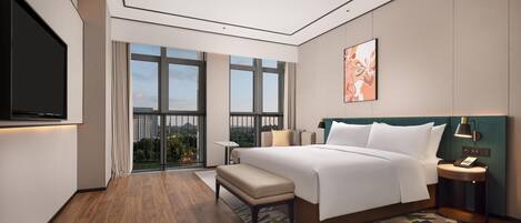 A modern hotel room with a large bed, a desk, a chair, a TV, and a balcony with a view of the city.