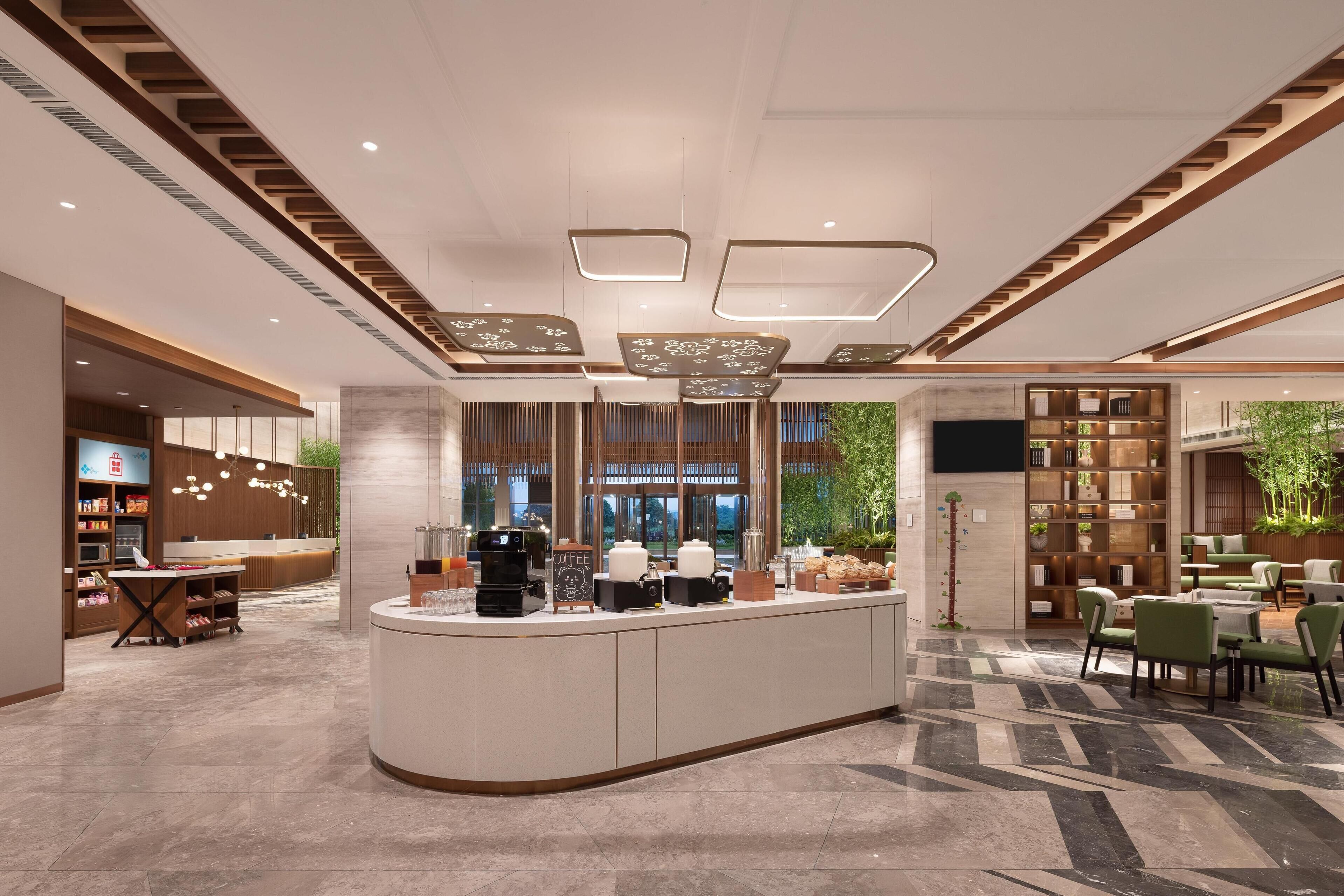 A modern hotel lobby with a central reception desk, seating area, and a large window offering a view of greenery.
