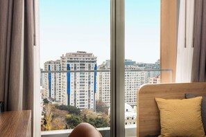 A modern hotel room with a large window offering a view of high-rise buildings and a park.