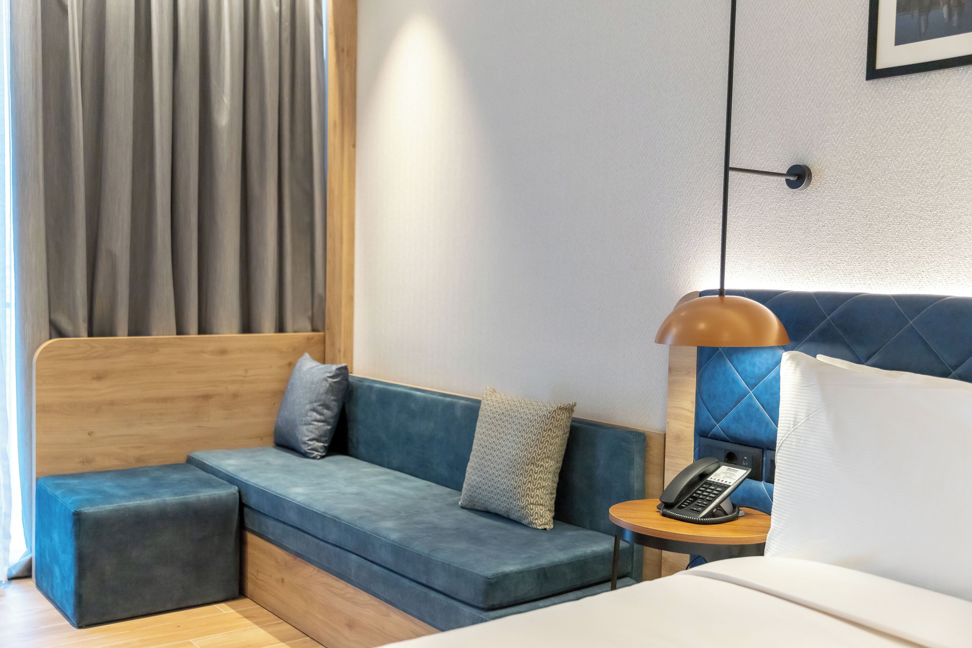 Ein modernes Hotelzimmer mit einem blauen Sofa, einem Holzkopfteil, einem Nachttisch mit Lampe und einem Telefon.