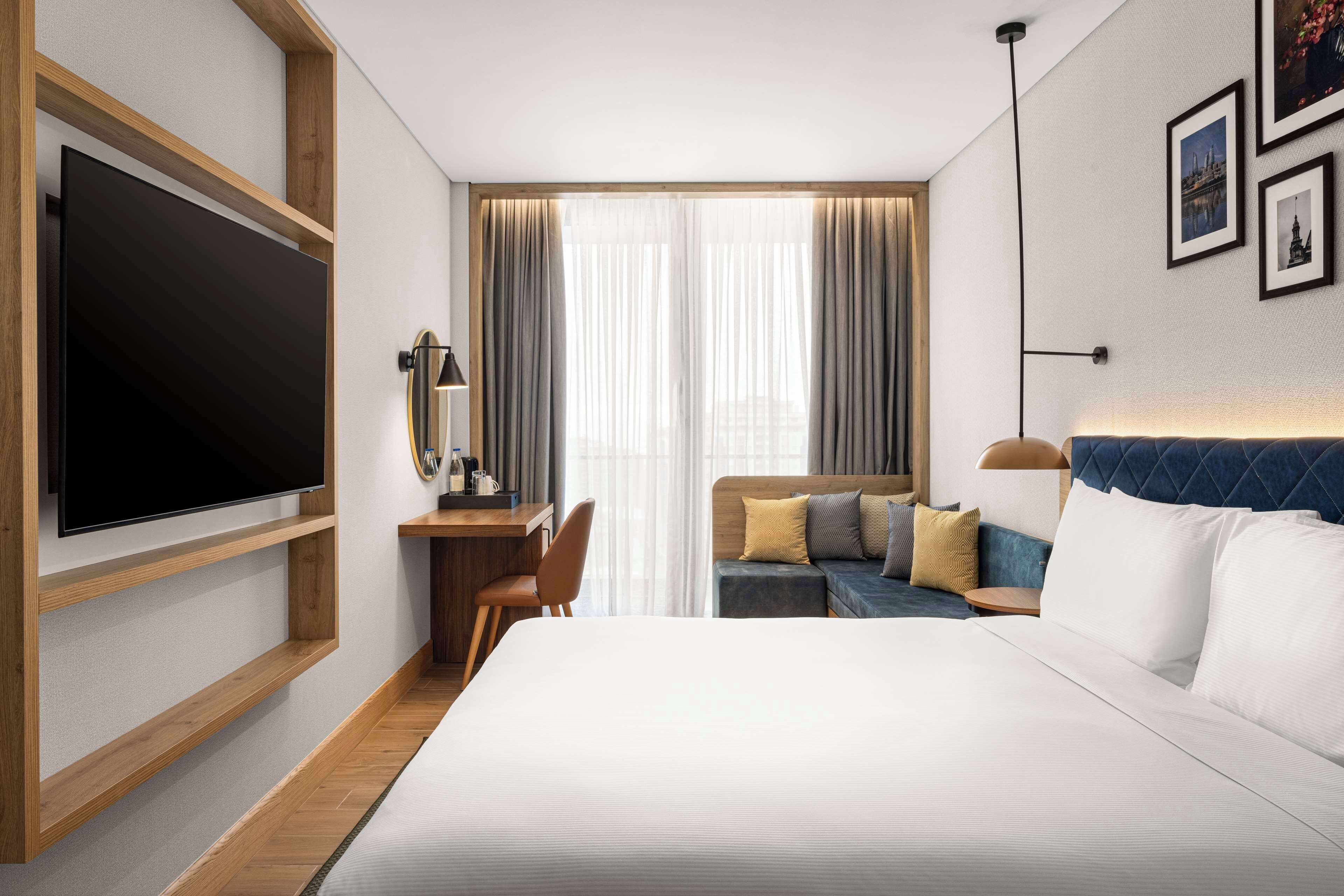 A modern hotel room with a large bed, a desk with a flat-screen TV, a comfortable sofa, and a large window with curtains.