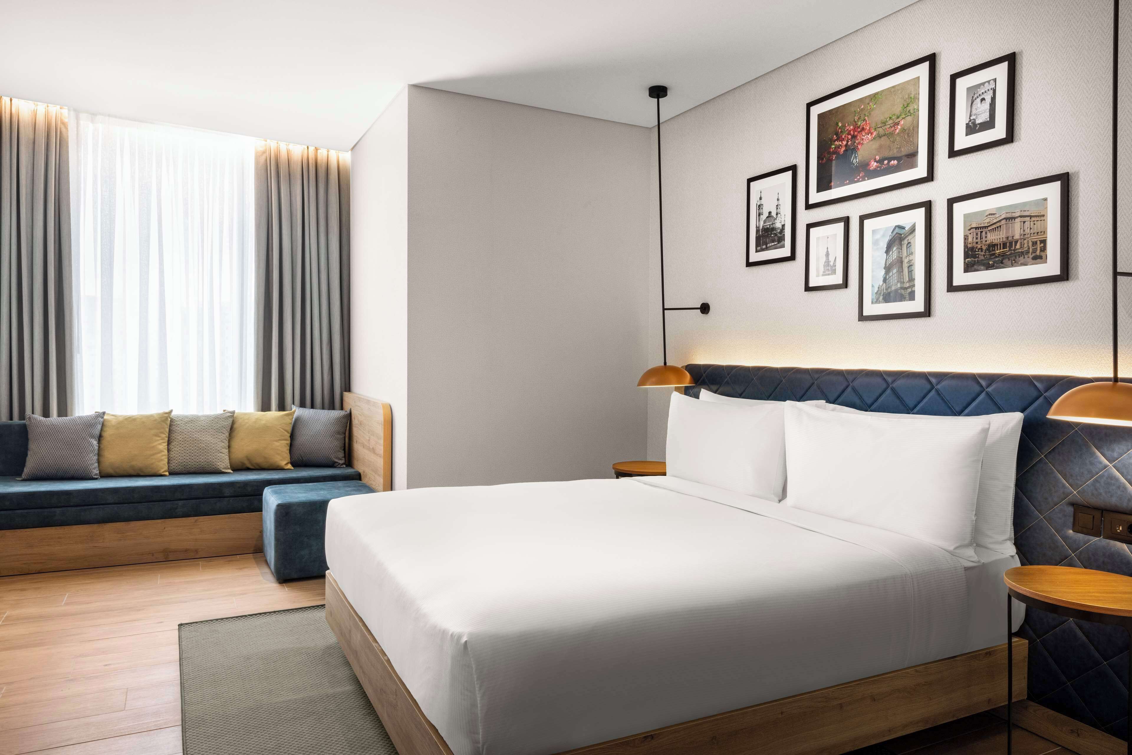 A modern hotel room with a large bed, a sofa, a small table, and a wall with framed pictures.